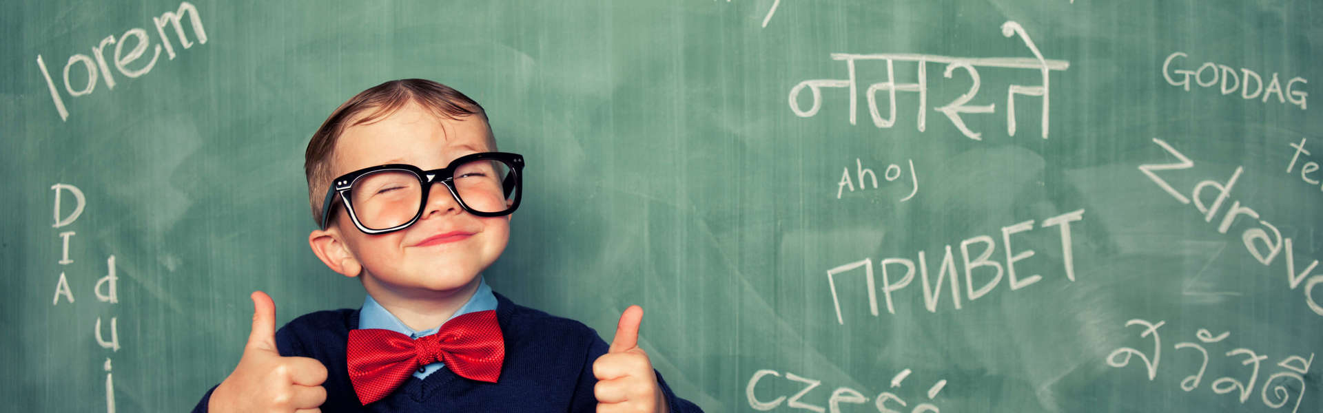 A young boy standing in front of a blackboard displaying 'hello' written in different languages